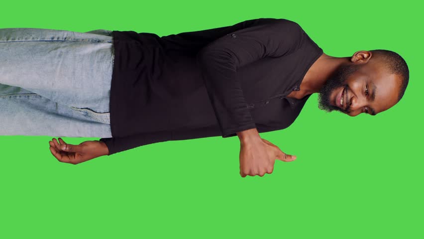 Vertical video: Side view of casual person giving thumbs up on camera, showing successful gesture and agreement. Young man doing like okay symbol with approval, standing over green screen background