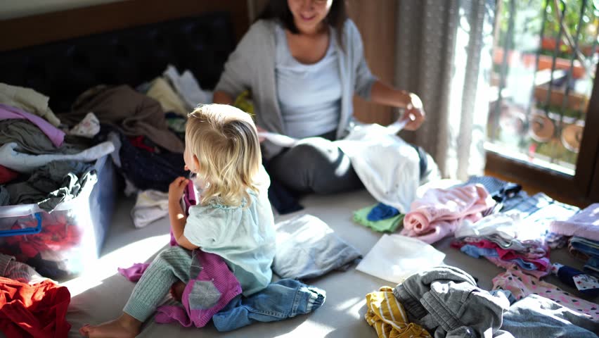 Little girl plays with clean linen that her mother folds on the bed
