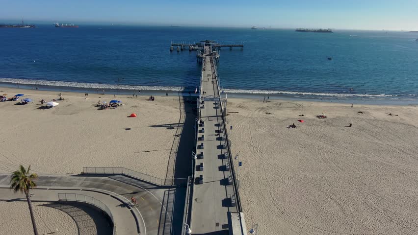 aerial footage of a concrete pier over the deep blue ocean water with people walking along the pier and curved light posts along the pier at Belmont Veterans Memorial Pier in Long Beach California USA