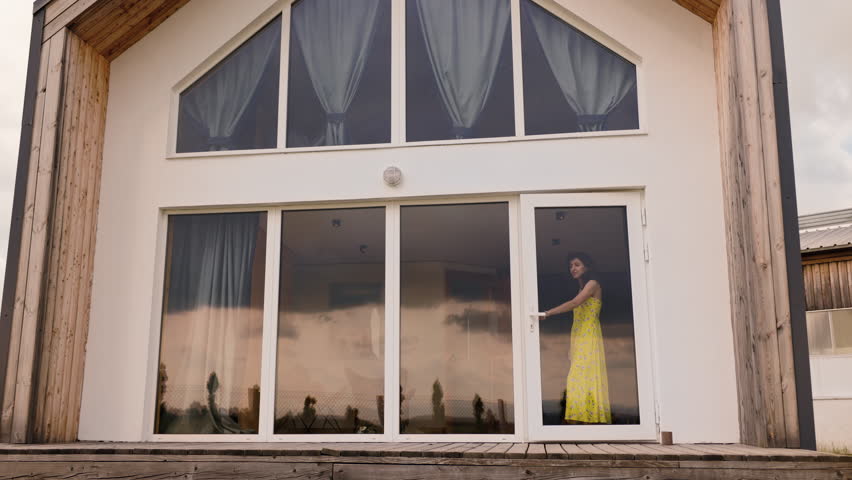 A woman wearing a yellow dress steps out of a house onto the porch barefoot. The house is a cozy barn-style building with a large terrace. The shot captures the full scene.