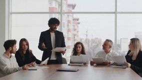 A young Indian businessman during a meeting explains to his partners the benefits of the company's new financial model. A group of people in business suits review reports and discuss ways to optimize
