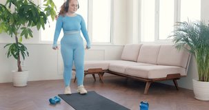 Cheerful young happy obese woman doing forward walking lunges, listening to music, wearing headphones, joining, keeping palms together