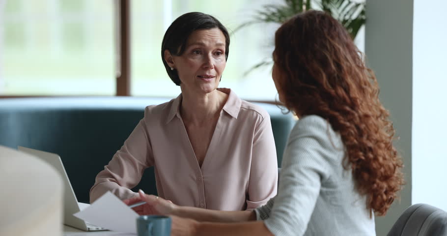 Mature business mentor woman talking to younger female employee, intern at workplace, speaking, teaching, explaining work tasks, asking questions, smiling, laughing, sitting at table with laptop Royalty-Free Stock Footage #1102351281