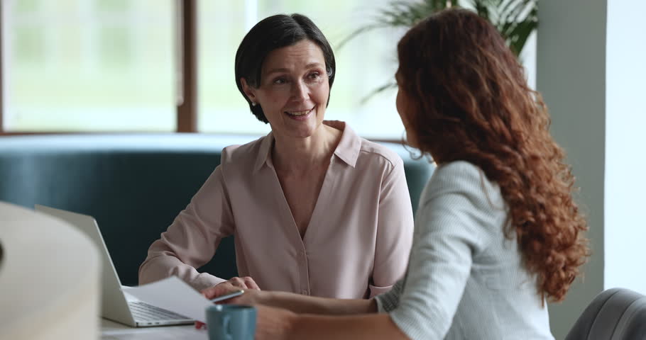 Mature business mentor woman talking to younger female employee, intern at workplace, speaking, teaching, explaining work tasks, asking questions, smiling, laughing, sitting at table with laptop