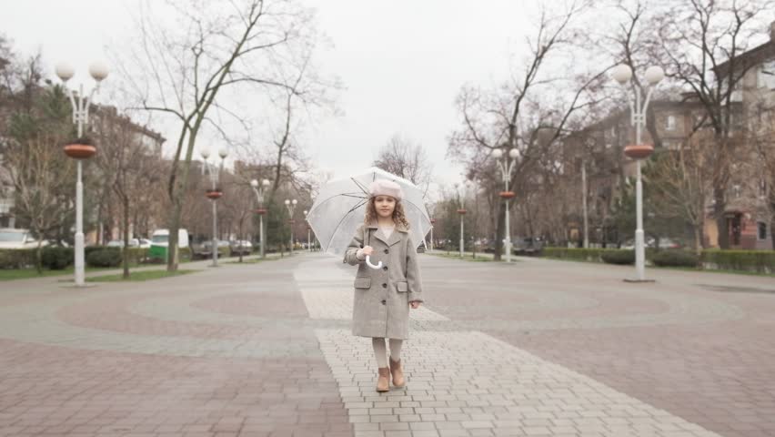 A beautiful little girl with a white umbrella walks along a city alley on a cloudy spring day.