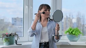 Middle aged woman doing makeup applying powder with brush looking in mirror - Powered by Shutterstock - Get 15% off with code: PIKWIZARD15