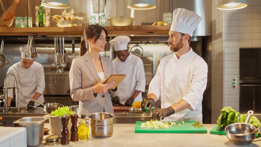 Portrait of attractive professional caucasian chef chopping vegetables and talking with beautiful cute female manager with tablet. Two colleagues discussing work at kitchen of restaurant. Royalty-Free Stock Footage #1102356745
