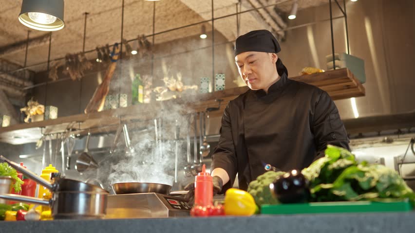Portrait of cheerful professional asian chef cooking food frying on pan with beautiful flames. Attractive skillful successful male cook wearing black uniform enjoying working at kitchen. Royalty-Free Stock Footage #1102356761