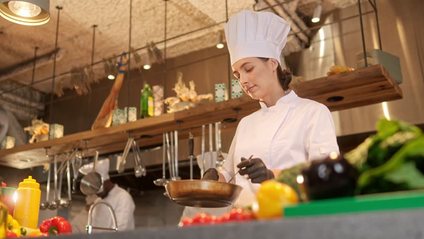 Attractive cute young caucasian female chef holding pan with delicious food tasting and smelling smiling. Beautiful professional chef cooking dishes in modern kitchen in white uniform.