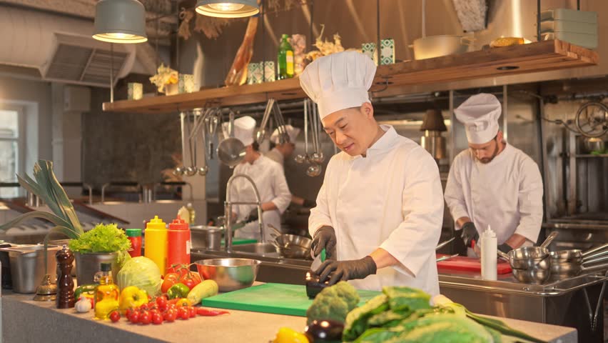Focused skillful professional Asian chef cooking delicious salad chopping aubergine on cutting board with knife. Concentrated male cook wearing white uniform and black gloves in kitchen.