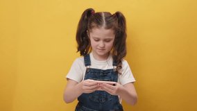 Portrait of funny girl kid enthusiastically playing racing or shooter video games on smartphone. Excited little child using mobile phone gadget app with drive simulator, isolated on yellow background