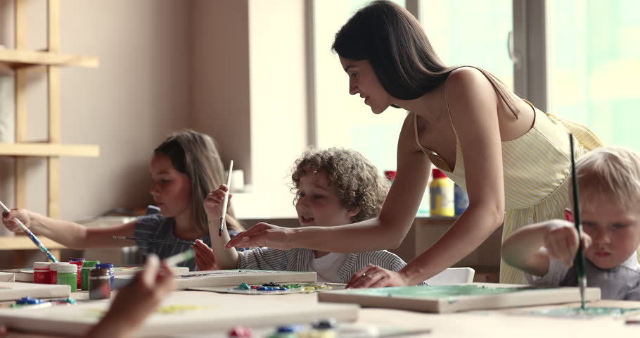Artistic school teacher talking to little pupil boy on class, helping with painting on canvas, checking artwork. Kid studying to draw in acrylic colors in studio, speaking to master woman Royalty-Free Stock Footage #1102361609