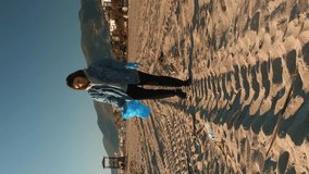 Girl in protective gloves collecting garbage on beach. Vertical video. Young woman cleaning plastic on beach. Volunteer collecting trash in trash bag. Plastic pollution