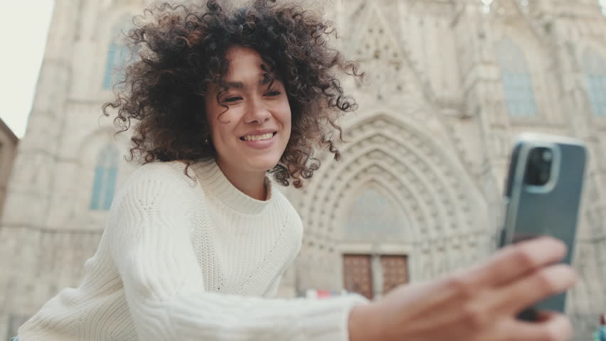 Young woman is sitting down on a cell phone. Girl showing victory sign posing for a selfie on the old city background