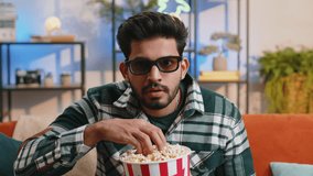 Excited indian man in 3D glasses sits on sofa eating popcorn snacks and watching interesting TV serial sport game, film, online social media movie content at home apartment. Guy enjoying entertainment