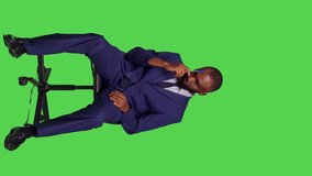 Vertical video: Front view of pensive employee thinking about solutions sitting on chair, posing over green screen wearing business office suit. Male entrepreneur brainstorming new ideas and feeling