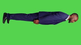 Vertical video: Profile of negative office worker doing thumbs down in studio, wearing formal suit over greenscreen background. Young businessman showing dislike gesture and expressing disapproval on