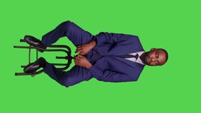 Vertical video: Front view of confident startup employee sitting on chair in studio, looking around and waiting for something. Young business manager wearing formal business suit over green screen
