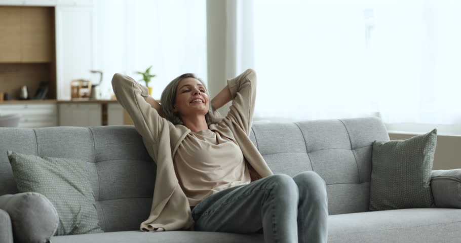 Cheerful middle aged woman walking to couch, sitting down, smiling, relaxing, stretching body with closed eyes, looking away in deep thoughts, thinking, dreaming