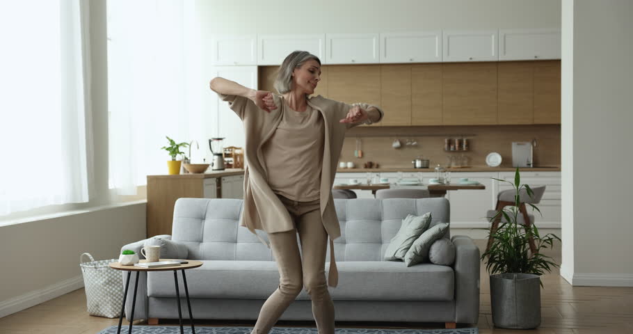 Positive active grey haired woman dancing at home, smiling, moving across living room, enjoying music, activity, movement, leisure, having fun. Mature lady celebrating success Royalty-Free Stock Footage #1102375243