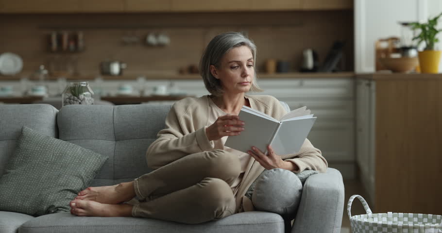 Focused middle aged woman relaxing at home, reading paper book, enjoying novel, leisure, studying new skills, getting knowledge, resting on comfortable couch at home Royalty-Free Stock Footage #1102375263