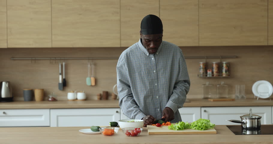 Happy African chef man in cook hat slicing vegetables for salad at kitchen table, dancing to music, preparing dinner from organic food products, natural ingredients, smiling, having fun