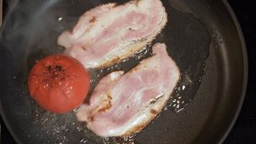Frying Bacon Slices in a Pan. Crispy Pieces of Delicious Red Thin Smokey Bacon Fried in a Hot Skillet with tomato. Traditional Breakfast. Fat High Calorie Food. Top view, Slow motion video