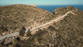 Aerial video of female cyclists in pink jersey descending on rural road on the hill with a sea view and Penon de Ifach rock in the background. Cycling in Calpe, Costa Blanca concept video.