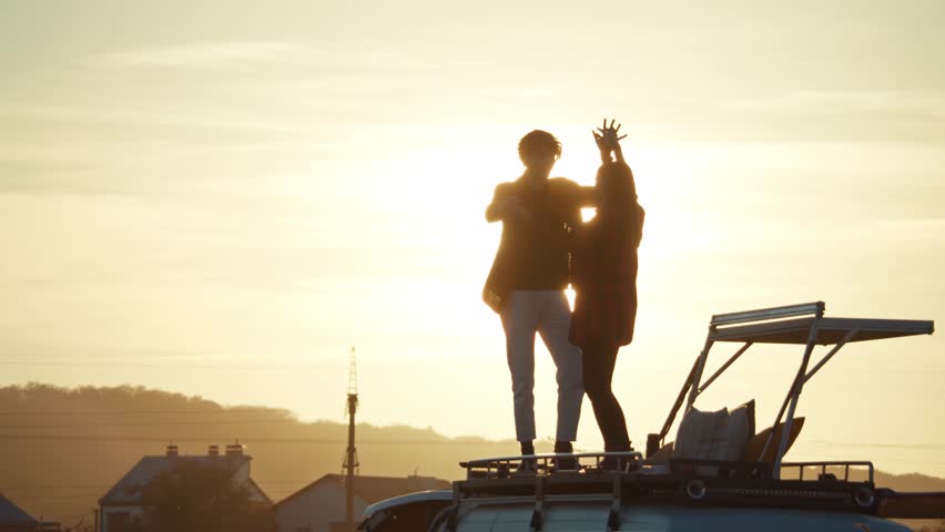 Beautiful view of hipster couple dancing on camper van rooftop, yellow sunset in background. Pair in love enjoying romantic road trip to backcountry. Dark silhouettes, yellow sky Royalty-Free Stock Footage #1102381367