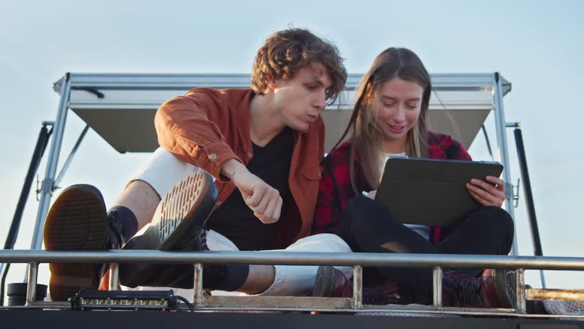 Young caucasian couple sitting on camper van rooftop, snacking on their road trip. Close-up, chewing hipster boy, girl using a tablet. Outdoor vacation, retreat to backcountry on weekend