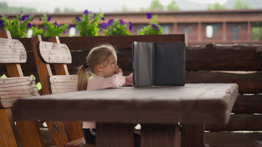Serious blonde little girl surfs internet on contemporary tablet computer sitting at large rustic table in hotel cafe on rainy day slow motion
