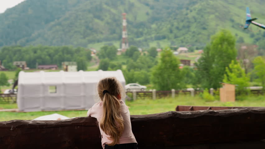 Curious little girl looks at flying up helicopter leaning on large wooden log railing on terrace of rural cafe on cloudy summer day slow motion