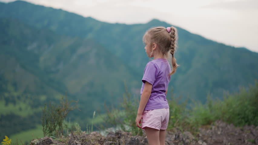 Cute little girl with braided hair looks at old distant mountain foggy silhouettes and scratches leg at highland backside view slow motion
