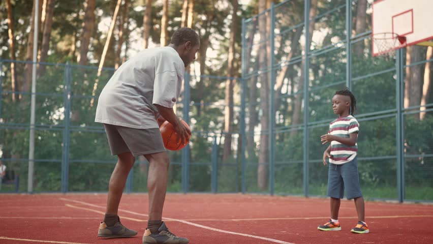 African American adult dad and little kid throwing each other ball playing basketball outside in city. Father and child play basketball on court in park. Sport and recreation. Family time together