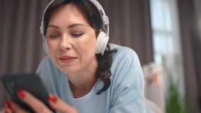 Pretty woman listening music through headphones with phone while lying on bed. Relaxing, wathing video clips of your favorite music. Close up. Musical device concept. Enjoying morning.