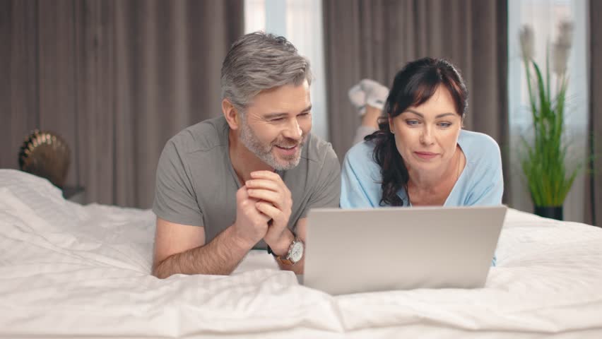 Attractive lovely woman and adult husband using computer on bed at home. Happy couple lying in bedroom having weekend, smiling, communicating and watching different videos together.