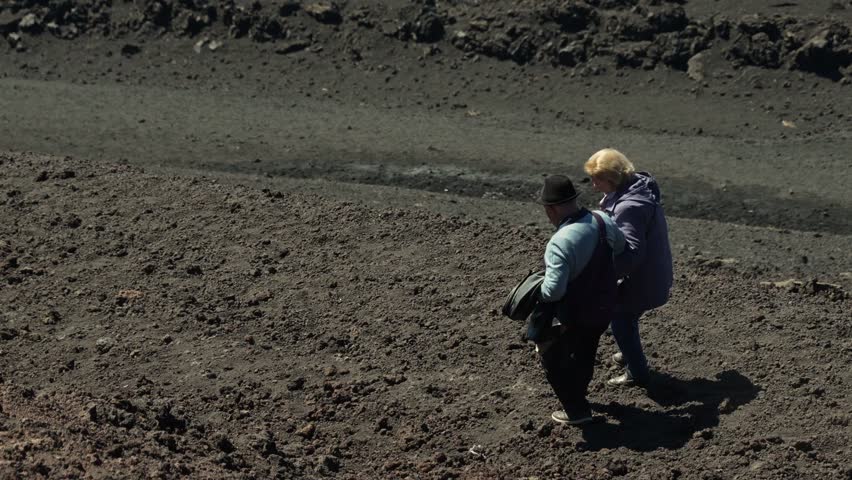 Elderly couple walking on lava ground. Old tourists hold hands descending a mountain. Hot air distortion, sun hits volcanic soil. Difficulty walking and standing. Love, affection, helping each other