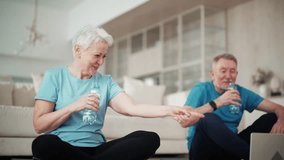 Happy senior couple gray haired man woman talk with fitness trainer in laptop after sport online training sit on mats at home in living room and drinks water. Aged family in sportswear after workout.