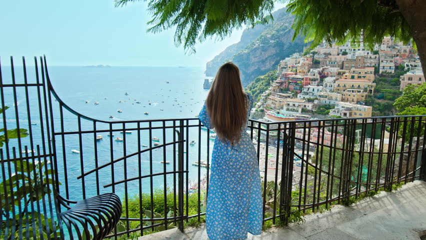 A female tourist exploring the cliffside of Positano. A young woman looking from above over the beautiful colorful houses and boats with the turquoise beach in Positano, Amalfi Coast. Royalty-Free Stock Footage #1102396191