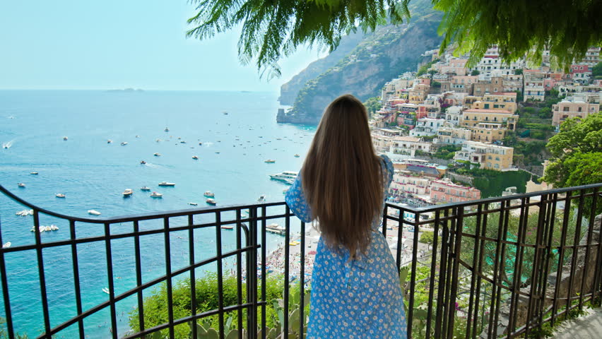 A female tourist exploring the cliffside of Positano. A young woman looking from above over the beautiful colorful houses and boats with the turquoise beach in Positano, Amalfi Coast.