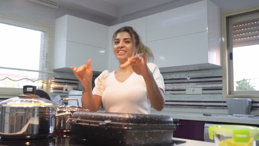 Middle Eastern cute happy woman dancing and singing while cooking dinner in kitchen at home. Frying burger, at pan domestic candid lifestyle routine.