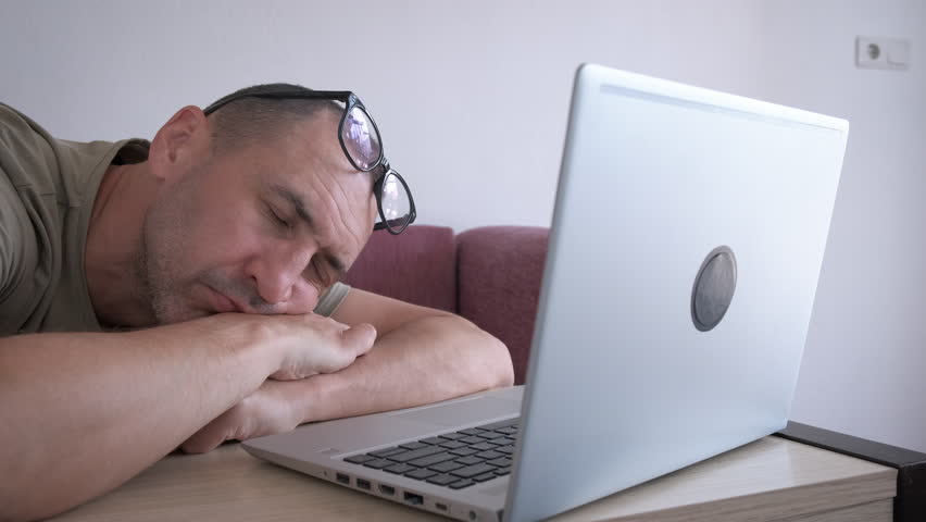 Man chronic sleep deprivation by computer. An exhausted man with chronic sleep deprivation sits by her computer in the room.