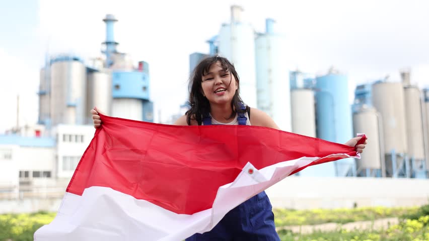 Cheerful asian female worker in hardhat with indonesian flag standing in front of factory Happy asian girl in work clothes and hardhat with flag of indonesia standing in front of industrial . High