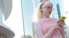 Pretty albino gen z teen girl holding smartphone device wearing earbuds using apps on cell phone listening mobile music looking at smartphone standing on city street lit with sunlight outdoors.