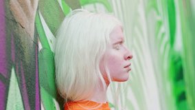 Pretty stylish albino teen generation z girl, cute inclusive beautiful female fashion model with white hair, pale skin standing at white wall with green leaves outdoors. Inclusion concept.