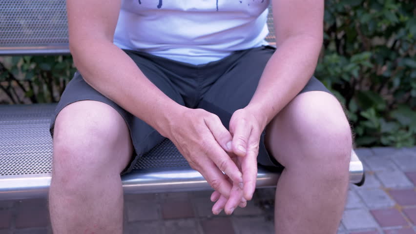 Male Rubbing Hands Sitting on a Bench in the Urban Park. A moment of waiting, excitement, anxiety, and emotional stress, trouble, nerves, and dread. Massage of hands, joints, fingers, skin. Outdoors.