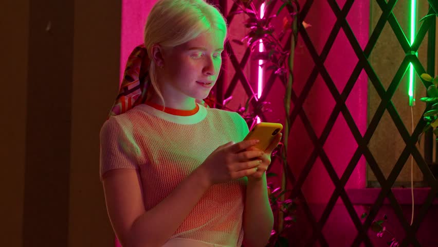 Pretty stylish albino gen z teen girl holding smartphone device typing text message using mobile apps on cell phone standing on city street lit with color neon night dark street outdoors.