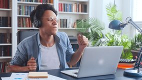 Young energetic attractive African American man millennial in headphones enjoys watching clips in laptops with youth music sits at table in home office with large bookcase. Rest, relaxation, lifestyle