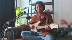 Young cute ethnic African American woman plays guitar in headphones and emotionally sings song about relationship with boyfriend or breaking up with lover sits in studio on couch near microphone