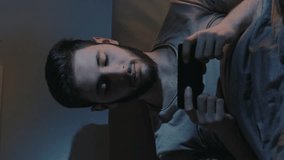Vertical video. Movie app. Gadget night. Online leisure. Tired sleepy man watching series on mobile phone late in bed in dark home bedroom.